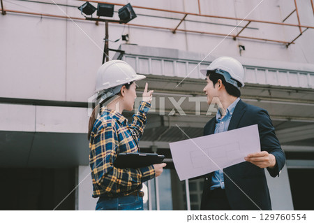 Construction manager and engineer dressed in orange work vests and hard helmets explore construction documentation on the building site near the steel frames Construction manager and engineer dressed in orange work vests and hard helmets explore construction documentation on the building site near the steel frames 129760554