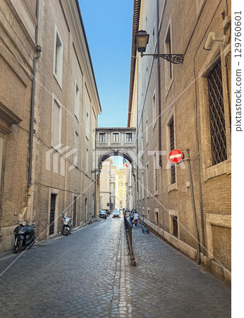 A long, narrow cobblestone street, lined with tall brick buildings and a unique overhead archway in Rome, Italy. Parked scooters and a few people walking the city A long, narrow cobblestone street, lined with tall brick buildings and a unique overhead archway in Rome, Italy. Parked scooters and a few people walking the city 129760601