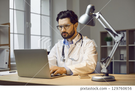 Portrait of serious, focused doctor sitting at his workplace at laptop in the process of filling out reports for the work done with patients. 129760656