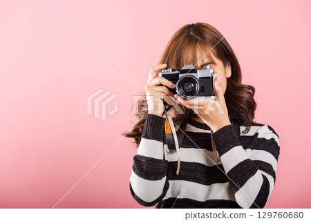Attractive energetic happy Asian portrait beautiful young woman smiling photographer taking a picture and looking viewfinder on retro vintage photo camera ready to shoot isolated on pink background 129760680