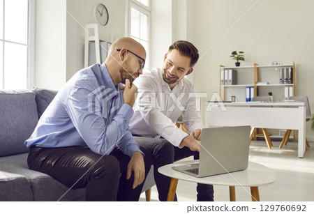 Two male colleagues using laptop together while sitting in office 129760692
