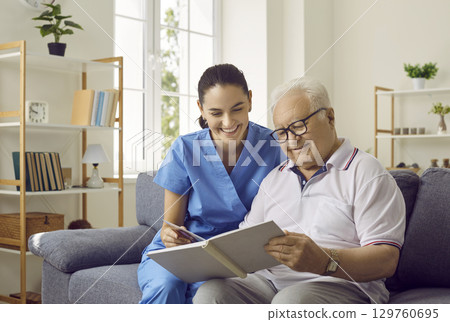 Friendly female nurse and older man are looking at photos from his family photo album together. 129760695