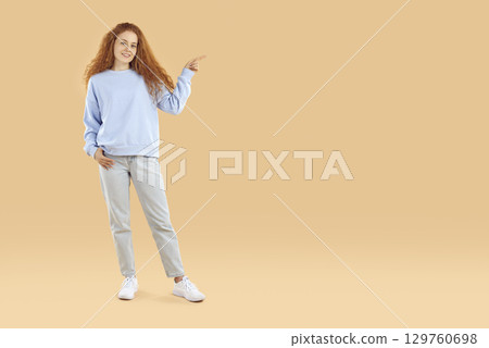 Young woman pointing to copy space and looking at camera with smiling face on a beige background. Young woman pointing to copy space and looking at camera with smiling face on a beige background. 129760698
