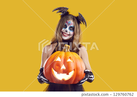 Happy girl in spooky Halloween costume with sugar skull makeup holding Jack-o-lantern Happy girl in spooky Halloween costume with sugar skull makeup holding Jack-o-lantern 129760762
