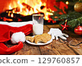 bitten oatmeal cookie and an unfinished glass of milk on a wooden table against the background of Santa Claus clothes a Christmas tree and a fireplace 129760857