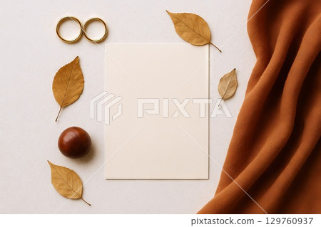 Minimalist autumn wedding flat lay with blank card surrounded by golden leaves, gold rings, chestnut and warm fabric. Concept of wedding invitation design, fall season celebration, romantic stationery 129760937