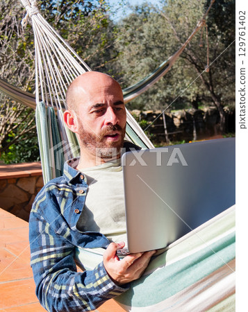 Smiling man working on laptop in hammock Smiling man working on laptop in hammock 129761402
