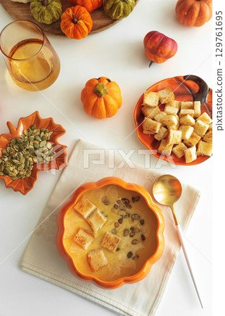 Creamy pumpkin soup with croutons and seeds, served in a pumpkin-shaped bowl on white wooden table. Top view Creamy pumpkin soup with croutons and seeds, served in a pumpkin-shaped bowl on white wooden table. Top view 129761695