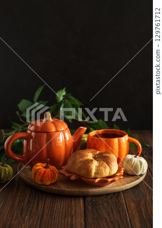 Autumnal tea set with pumpkin-shaped teapot and mug, and a baked bun on a wooden table. 129761712