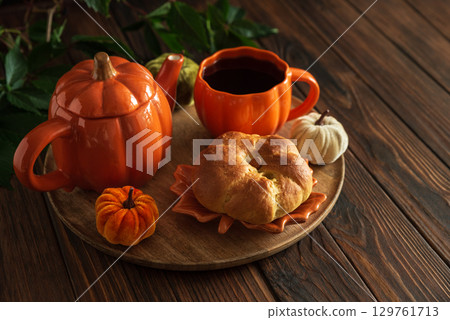 Autumnal tea set with pumpkin-shaped teapot and mug, and a baked bun on a wooden table. 129761713