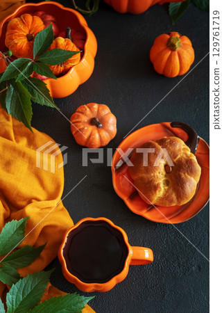 Autumnal tea set with pumpkin-shaped teapot and mug, and a baked bun on a black wooden table. 129761719