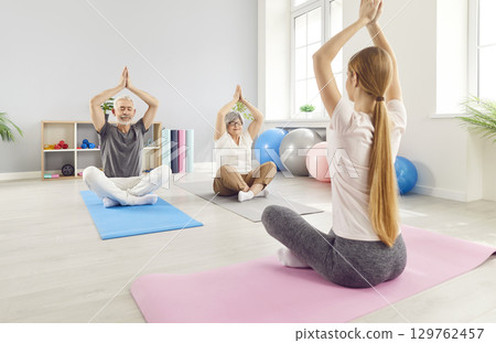 Mature senior people sitting on the floor on mat and practicing yoga in gym in Lotus pose Mature senior people sitting on the floor on mat and practicing yoga in gym in Lotus pose 129762457