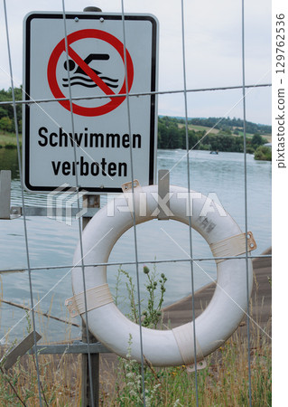 No swimming sign with a safety ring by a river, emphasizing caution. The sign prohibits swimming to ensure safety near the water. Vegetation surrounds the sign No swimming sign with a safety ring by a river, emphasizing caution. The sign prohibits swimming to ensure safety near the water. Vegetation surrounds the sign 129762536