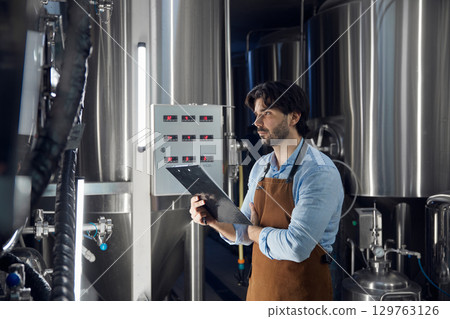 A man brewer in an apron stands confidently in a factory, arms crossed A man brewer in an apron stands confidently in a factory, arms crossed 129763126