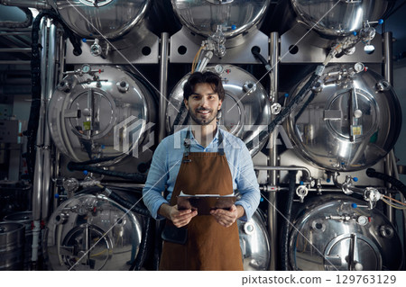 A man wearing an apron is holding a tablet inside a brewery facility A man wearing an apron is holding a tablet inside a brewery facility 129763129