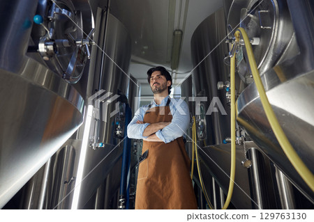 A man wearing an apron is standing with arms crossed in a factory 129763130