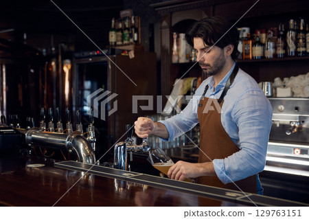 A bartender skillfully pours a beer from a tap into a glass container 129763151