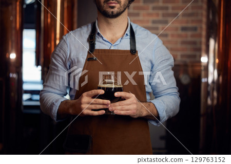 A man wearing an apron is holding a frosty glass of beer 129763152