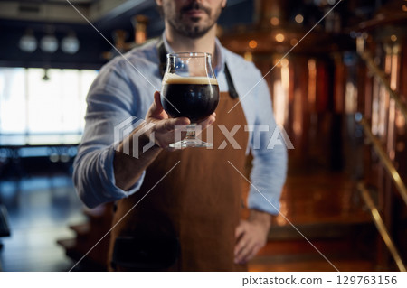 A man brewer wearing an apron and is holding a glass filled with beer A man brewer wearing an apron and is holding a glass filled with beer 129763156