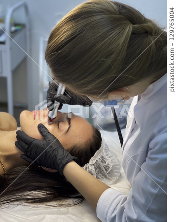 The cosmetologist in medical gloves doing a hydra peeling procedure on the client's cheeks. Side view. The cosmetologist in medical gloves doing a hydra peeling procedure on the client's cheeks. Side view. 129765004