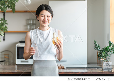 Young Asian woman in sportswear eating a banana in the kitchen (training, energy replenishment, snack) 129767040