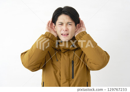 一位身穿羽絨外套的男子用手摀住耳朵聆聽，白色背景 129767233