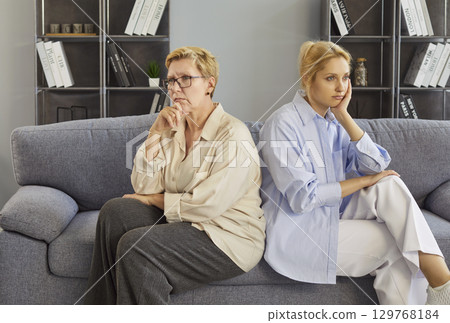Mother and daughter home conflict, sitting back-to-back on sofa, senior adult and young woman upset 129768184