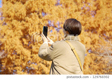 Woman taking a photo of autumn leaves, middle-aged, back view 129768302