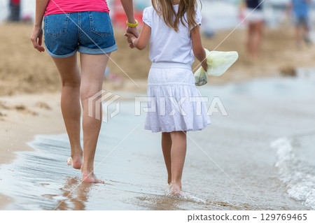 Mother and daughter girl walking together on sand beach in sea water in summer with bare feet in warm ocean waves. 129769465