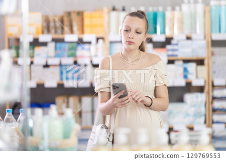 Young pregnant woman scrolling phone while looking for something in drugstore Young pregnant woman scrolling phone while looking for something in drugstore 129769553