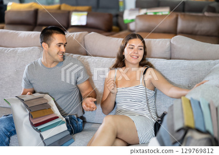 In store wife sit with legs on gray sofa to test in use,husband examines upholstery fabric 129769812