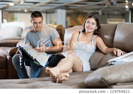 In store wife settled down with legs to test of sofa in use,husband examines upholstery fabric 129769821