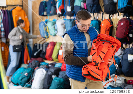 Middle-aged man meticulously chooses backpack for winter hike in travel equipment store 129769838