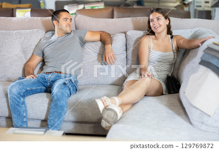 In store,husband and wife are sitting on gray sofa and choose upholstered furniture for living room 129769872