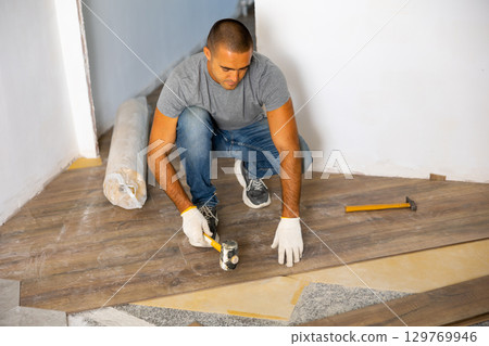 Concentrated man laying laminate flooring, strikes with soft hammer on part with lock for fixing. Concept of repair in house Concentrated man laying laminate flooring, strikes with soft hammer on part with lock for fixing. Concept of repair in house 129769946