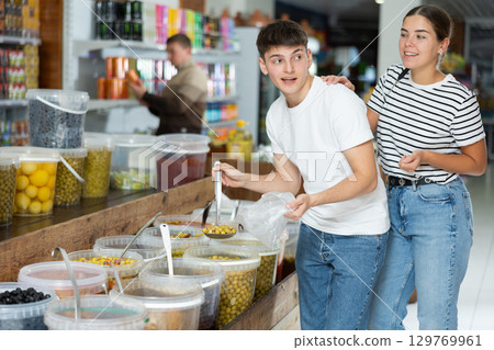 Young couple chooses marinated olives in supermarket, scooping them from bucket with ladle into plastic bag Young couple chooses marinated olives in supermarket, scooping them from bucket with ladle into plastic bag 129769961