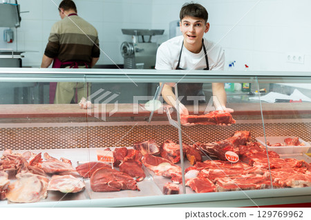 Young guy stands behind counter in meat department of supermarket and and holds piece of raw meat in his hands Young guy stands behind counter in meat department of supermarket and and holds piece of raw meat in his hands 129769962