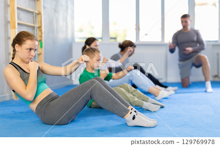 Concentrated girl, boy, woman, man at group training with trainer in gym practicing strikes in sitting position Concentrated girl, boy, woman, man at group training with trainer in gym practicing strikes in sitting position 129769978