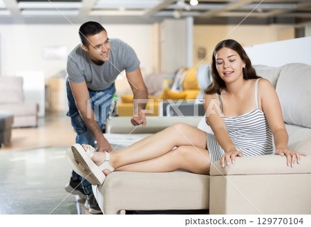 Couple chooses furniture in store.Wife is sitting on sofa,husband is standing next to her 129770104