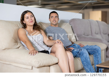 In store,husband and wife are sitting on gray sofa and choose upholstered furniture for living room 129770155