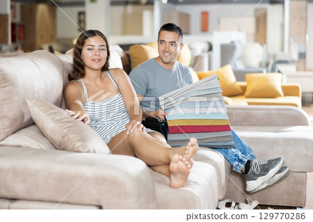 In store wife sit with legs on gray sofa to test in use,husband examines upholstery fabric 129770286