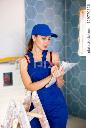 Woman in blue overalls checking completed construction work on drawing Woman in blue overalls checking completed construction work on drawing 129770326