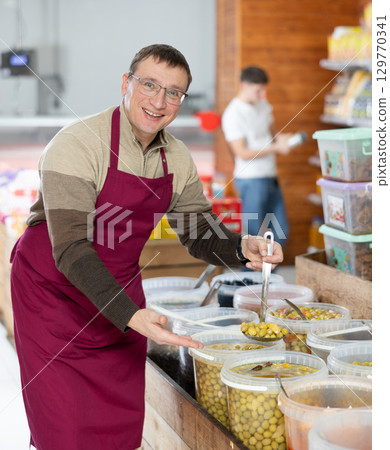 Seller man in supermarket showing and scooping marinated green olives 129770341