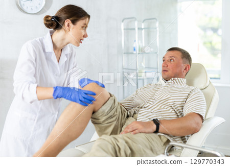 Female doctor carefully examining the sore leg of a male patient in clinic 129770343