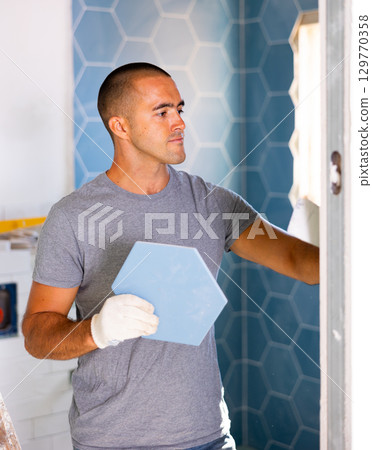 Portrait of concentrated young man in ordinary clothes installing ceramic tiles in bathroom Portrait of concentrated young man in ordinary clothes installing ceramic tiles in bathroom 129770358