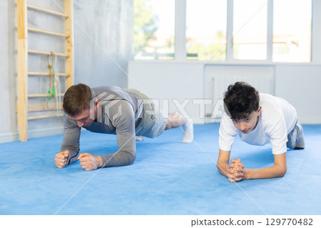 Underage boy and middle-aged trainer practicing plank pose in sports hall 129770482