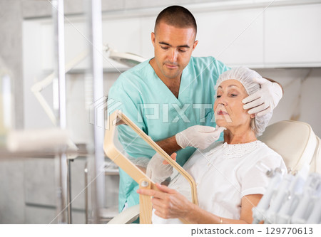 Male plastic surgeon giving professional advice after facial contouring to senior woman client looking at mirror Male plastic surgeon giving professional advice after facial contouring to senior woman client looking at mirror 129770613