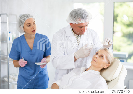 Elderly woman getting procedure of injection contouring and for facial correction in cosmetology clinic Elderly woman getting procedure of injection contouring and for facial correction in cosmetology clinic 129770700