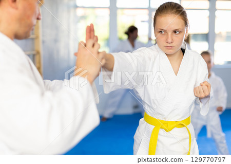 Teen girl at group training in gym practice karate punch block technique with trainer Teen girl at group training in gym practice karate punch block technique with trainer 129770797