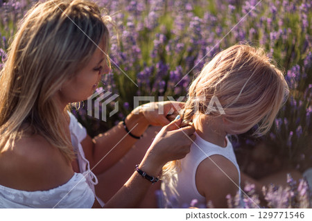 Mother Daughter Lavender: Woman braids girl's hair amidst purple flowers during a sunny day bonding. 129771546